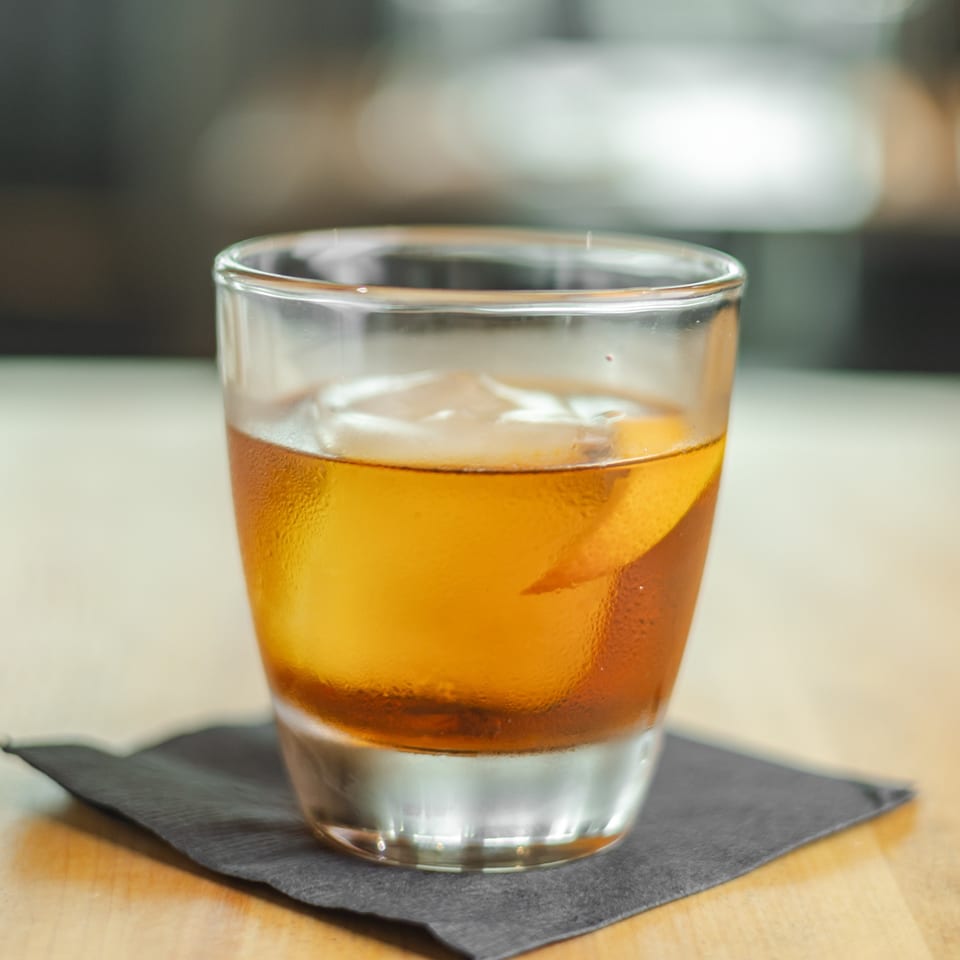 Amber cocktail with ice and an orange peel garnish served in a tumbler on a black slate coaster.