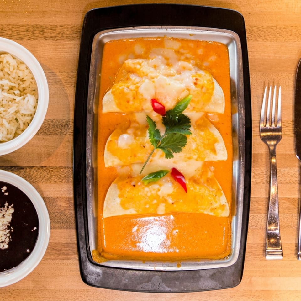 Three enchiladas covered in orange sauce, garnished with cilantro and peppers, served with rice and black beans on a wooden table.