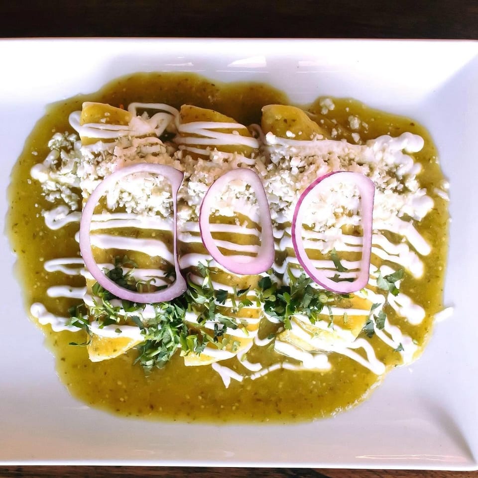 Enchiladas smothered in green sauce, drizzled with crema, topped with crumbled cheese, red onion rings, and cilantro.