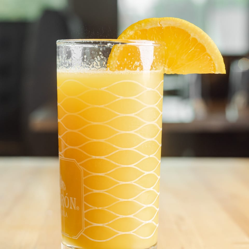Bright orange beverage in a patterned glass, garnished with a fresh orange slice, sits on a restaurant table.