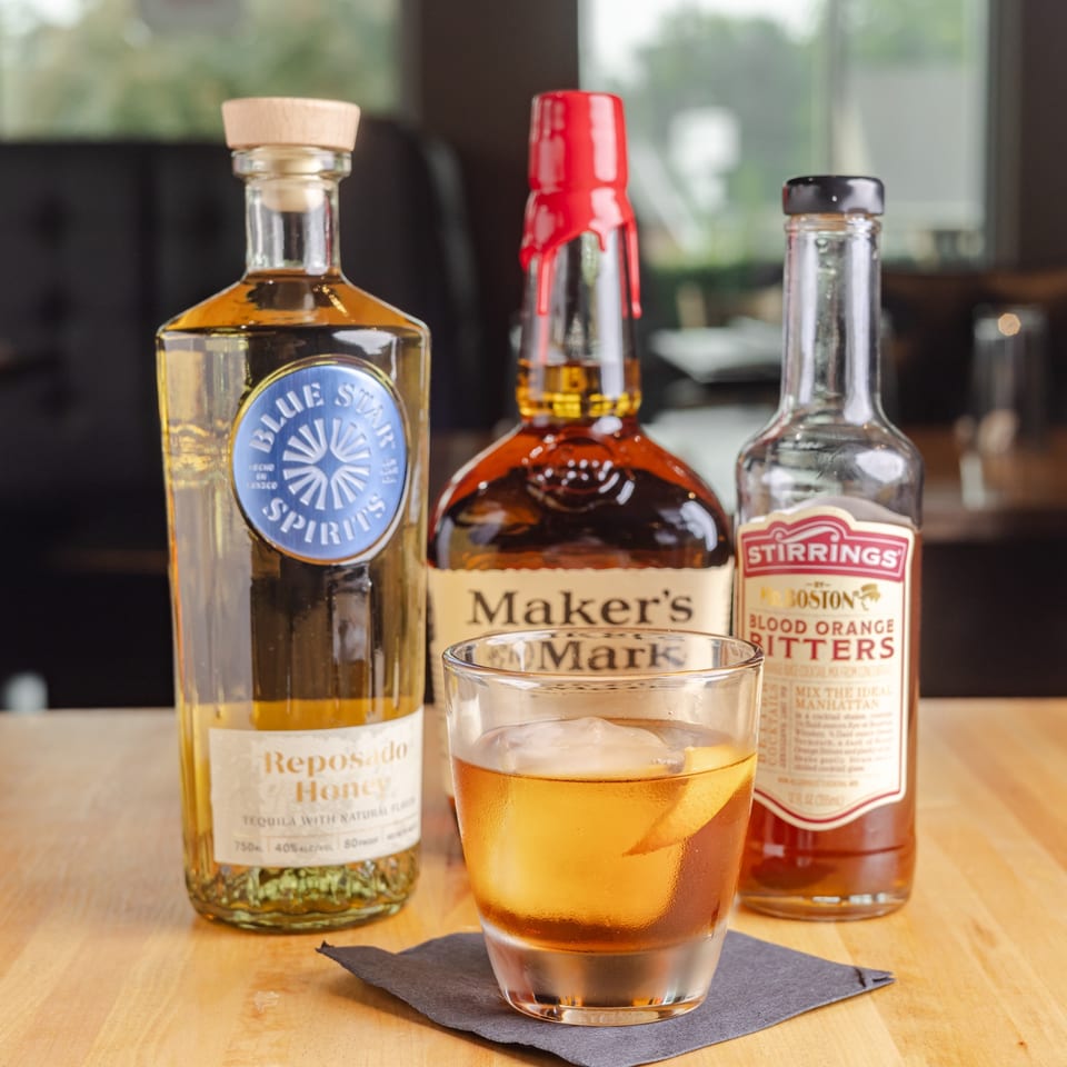 Amber cocktail with ice and citrus peel sits on a napkin beside tequila, bourbon, and blood orange bitters bottles.
