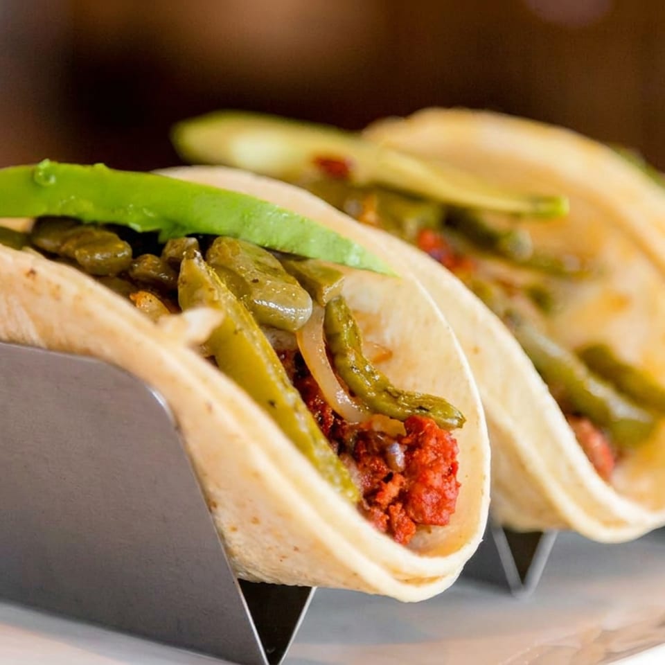 Three soft tacos filled with seasoned meat, sautéed peppers, onions, and avocado arranged in a metal holder.