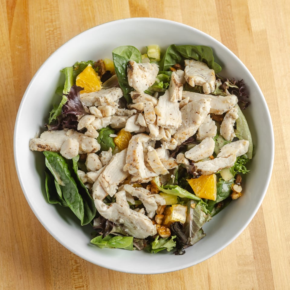 Top-down view of a salad bowl with grilled chicken, mixed greens, orange segments, cucumber, avocado, and nuts.