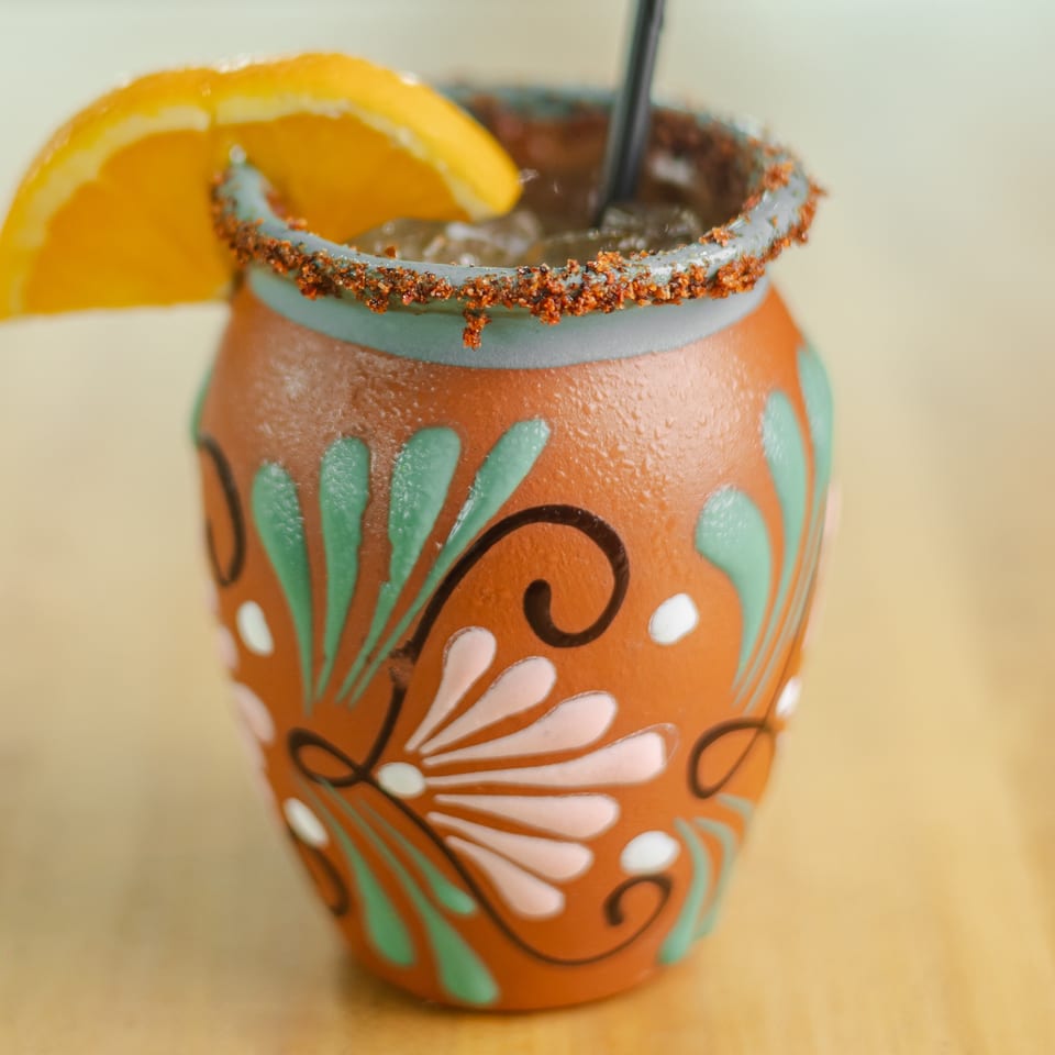 Close-up of an orange cocktail in a colorful floral ceramic glass, rimmed with chili seasoning and garnished with an orange slice.