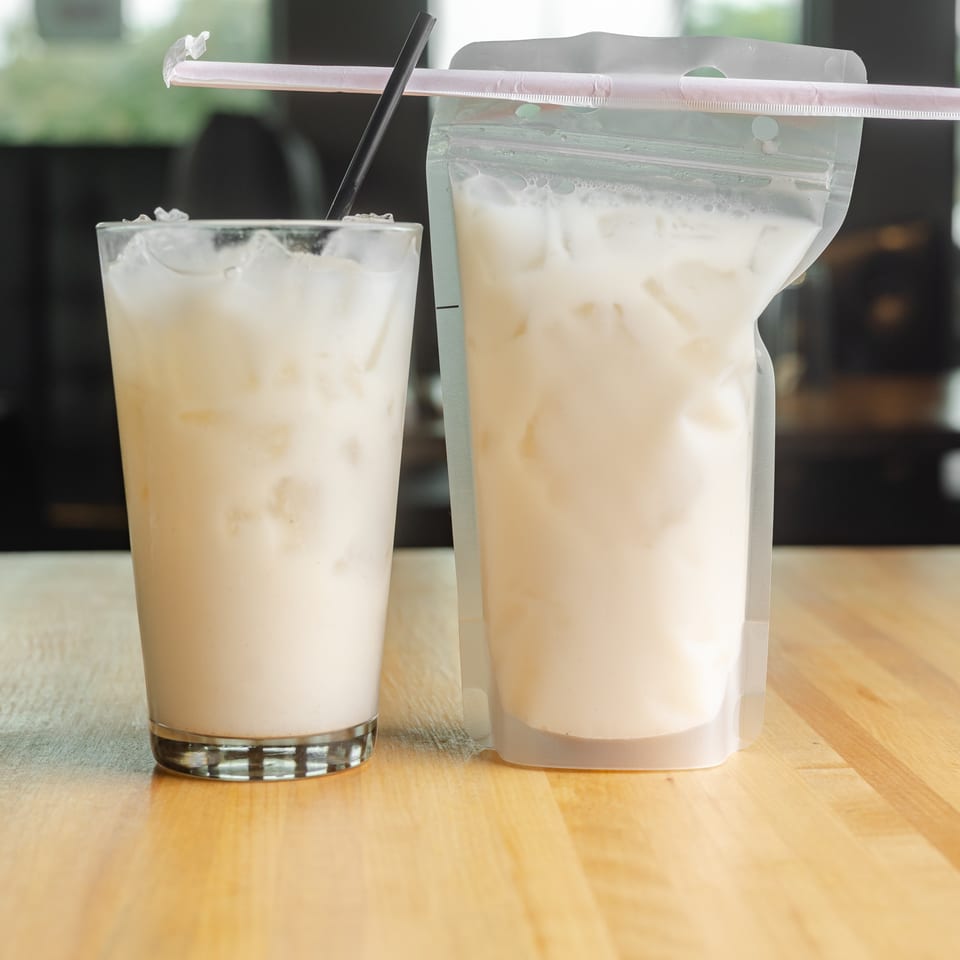 Two creamy iced beverages sit on a wooden restaurant table, one in a glass and the other in a resealable pouch.