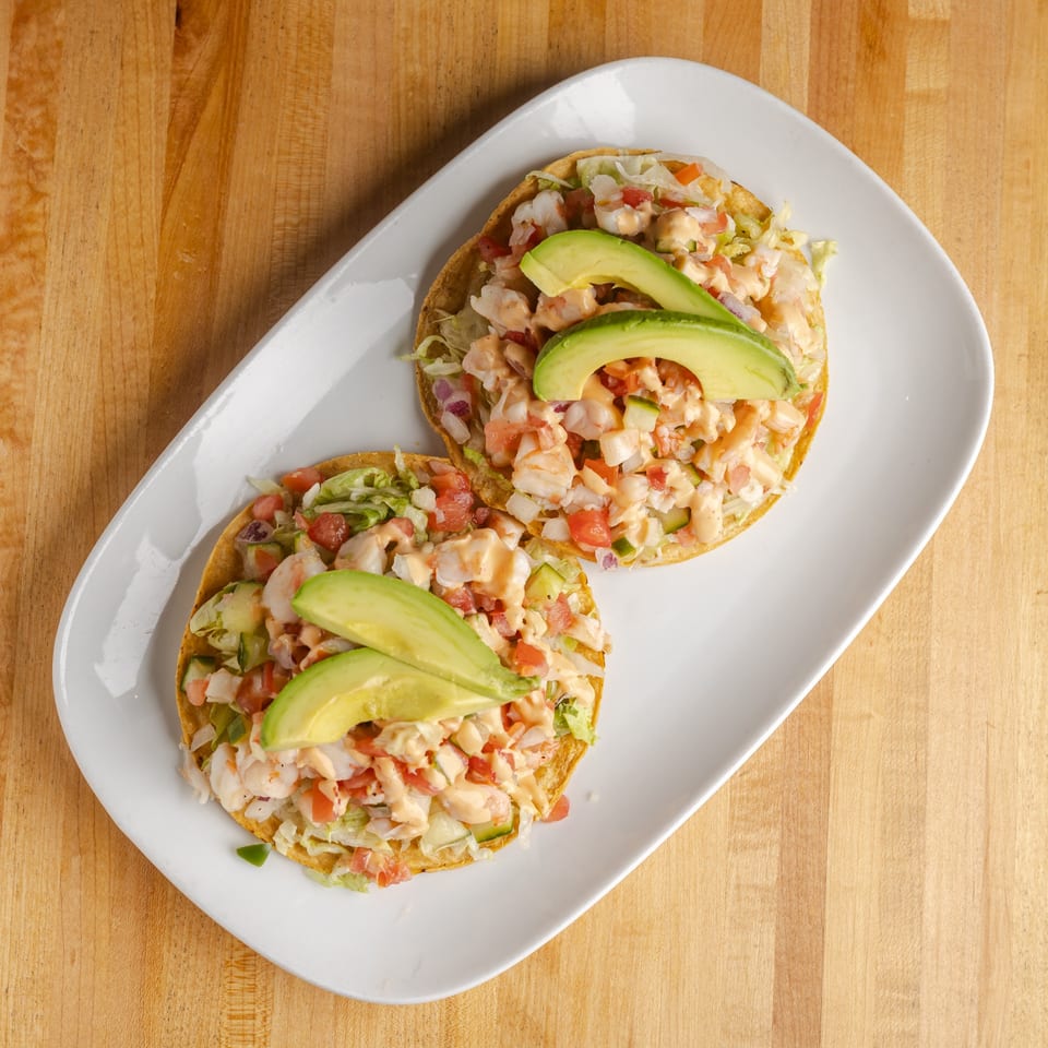Two shrimp tostadas topped with lettuce, pico de gallo, creamy sauce, and avocado slices on a white plate.